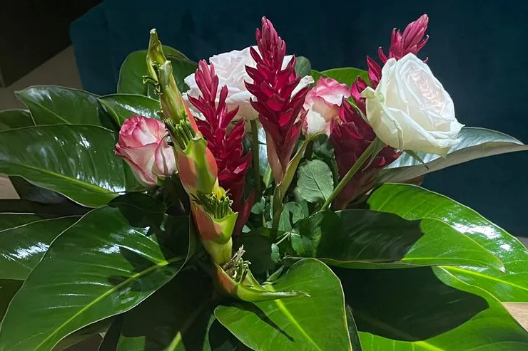 An arrangement of red ginger, white and pink roses in a basket, ringed with broad glossy leaves